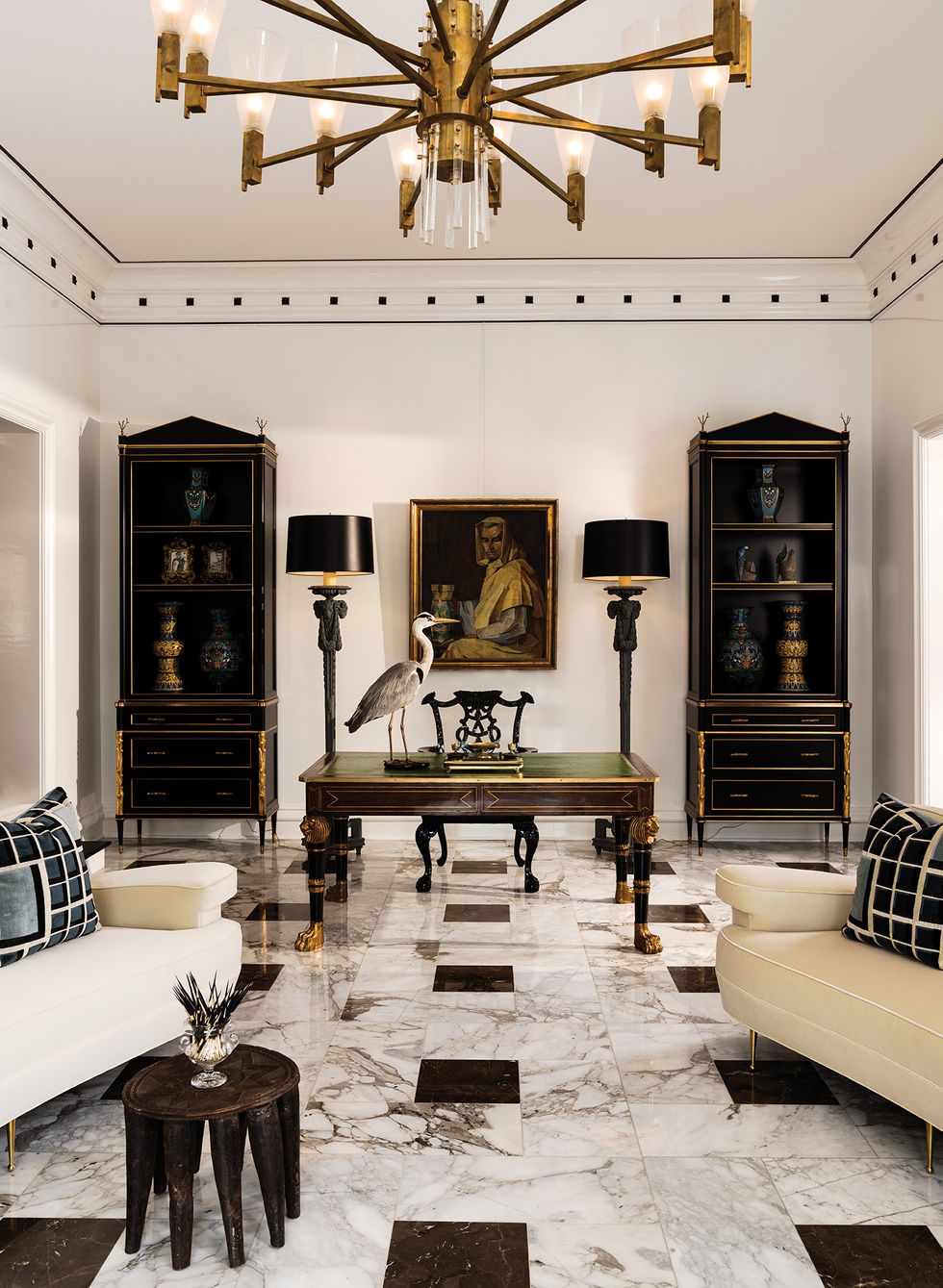 An Empire-style desk is flanked by ebonized Neoclassical bookcases, while a vintage 16-arm brass chandelier hangs above