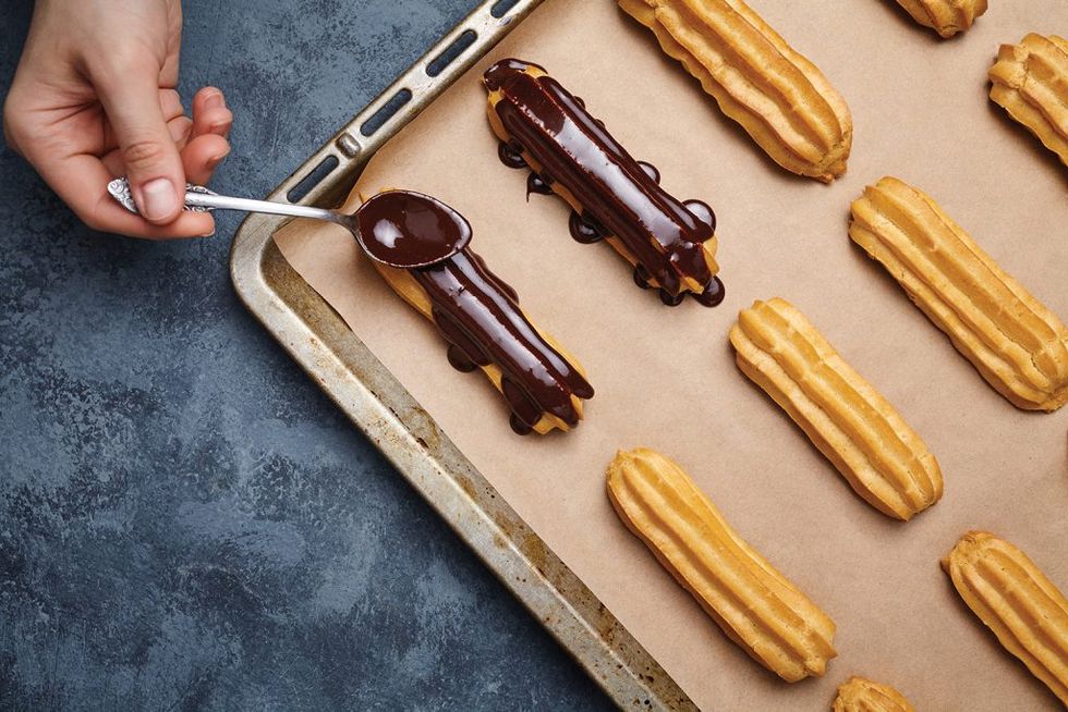 Eclairs or profiterole with chocolate and whipped cream preparing on baking sheet. Spreading chocolate on top of eclair with spoon. Traditional French dessert. Empty space for design text template.